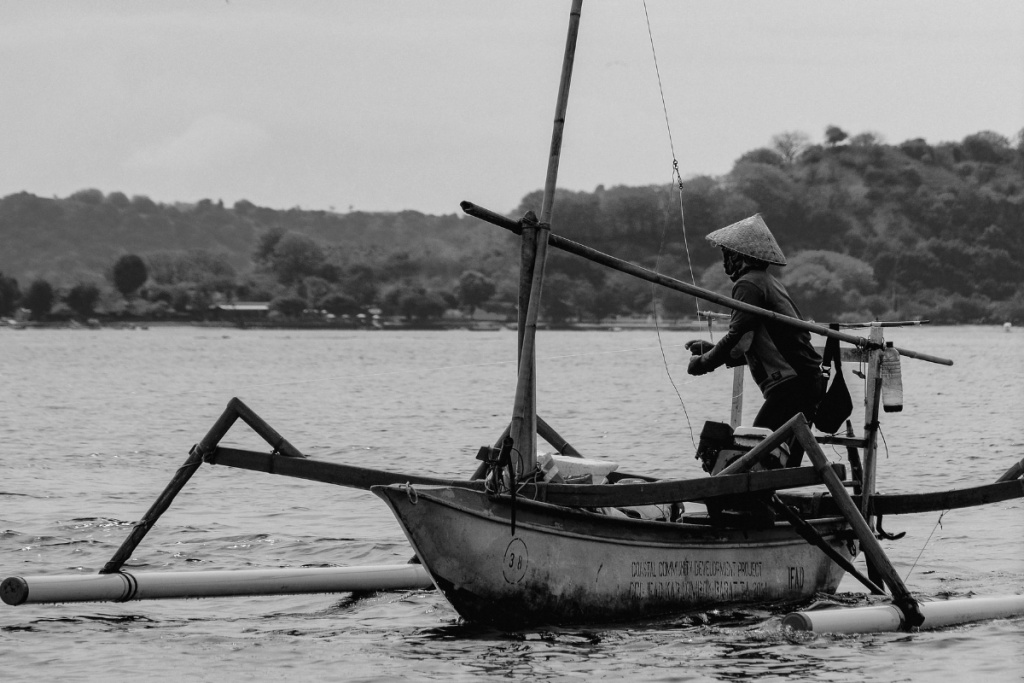 A fisherman wearing a traditional conical hat aboard a wooden outrigger boat in Lombok, Indonesia.