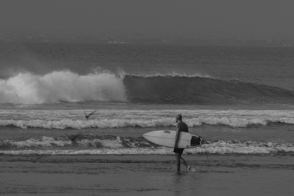 A surfer carrying a surfboard along the beach as a large wave breaks in Uluwatu, Bali.