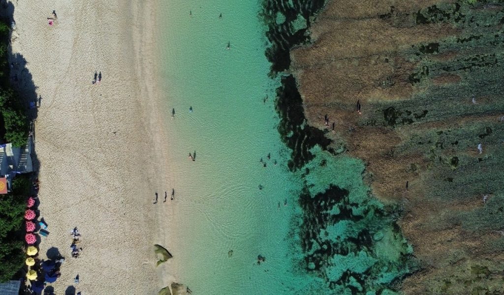 Aerial drone view of Melasti Beach in Uluwatu, Bali, with turquoise waters and white sand.