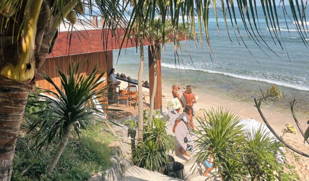 Surfer walking along Nyang Nyang Beach in Uluwatu, Bali, with board under arm and waves in the background.