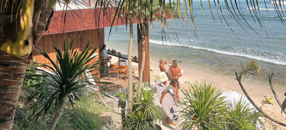 Surfer walking along Nyang Nyang Beach in Uluwatu, Bali, with board under arm and waves in the background.